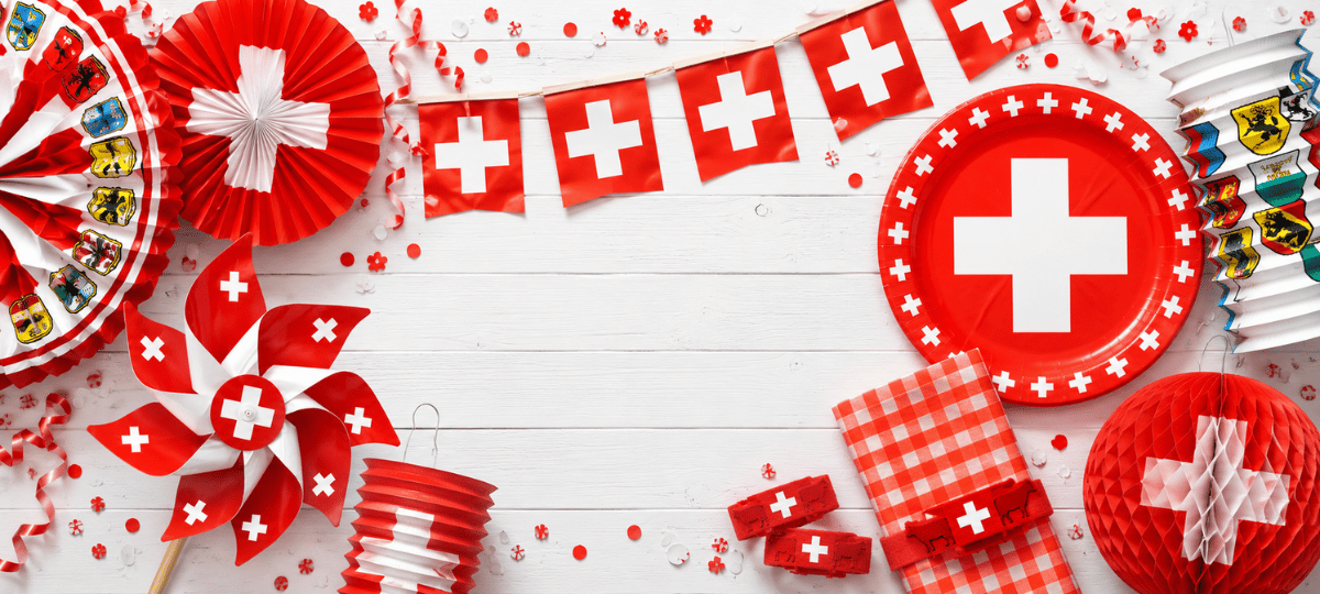 Schweizer 1.-August-Dekoration mit Papierfächern, Lampions, Fähnchengirlande, Windrad, Pappteller und Servietten in Rot-Weiss auf hellem Holzhintergrund – festlicher Banner für Nationalfeiertag und Sommerfeier.